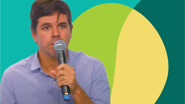 Foto recortada de homem branco de olhos claros falando ao microfone.